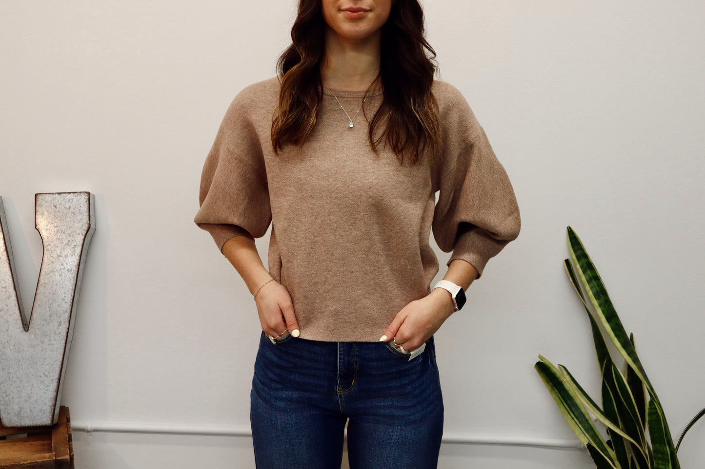 Woman wearing a brown sweater and blue jeans standing indoors with a plant and decorative letter in the background.