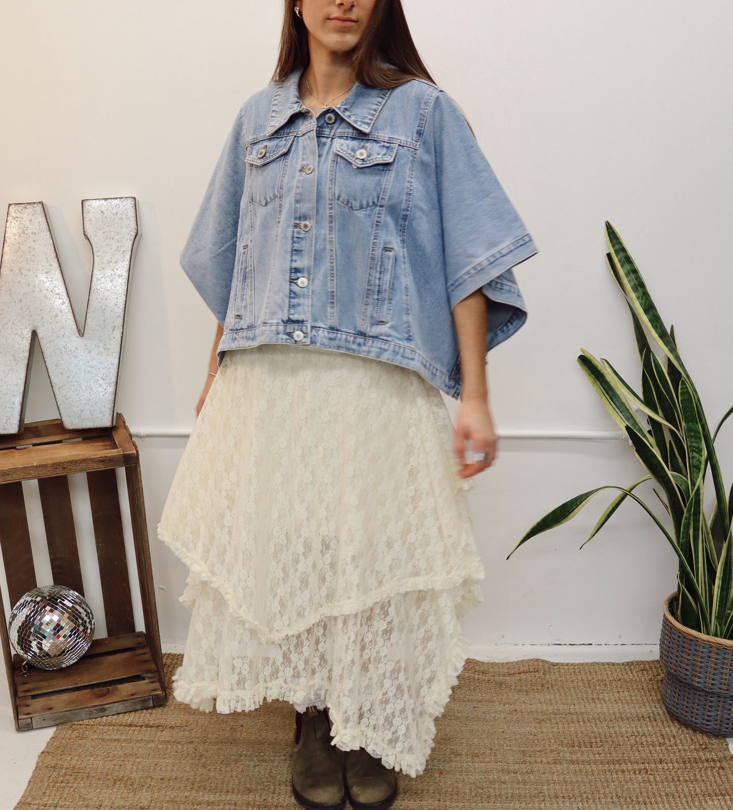 Person wearing a white sweater and blue denim jacket in a minimalistic room with a plant and wooden crate.