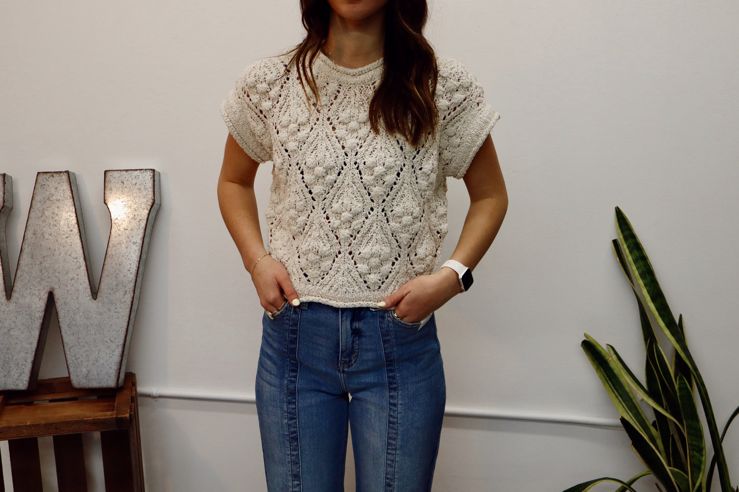 Person wearing a textured beige top and blue jeans standing indoors with decorative elements in the background.
